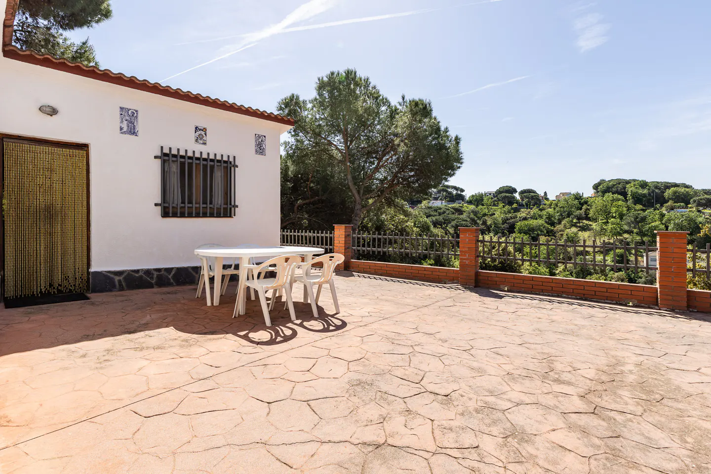 Outdoor patio with white table and chairs. A white building with a beaded door is on the left. A brick and metal fence overlooks green trees.