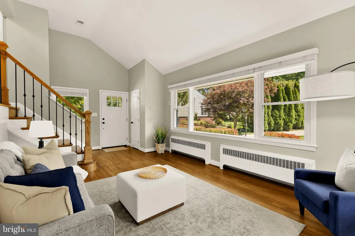 Living room with hardwood floors, gray walls, and large windows. A staircase is on the left, and a gray sofa with blue pillows is in the foreground.