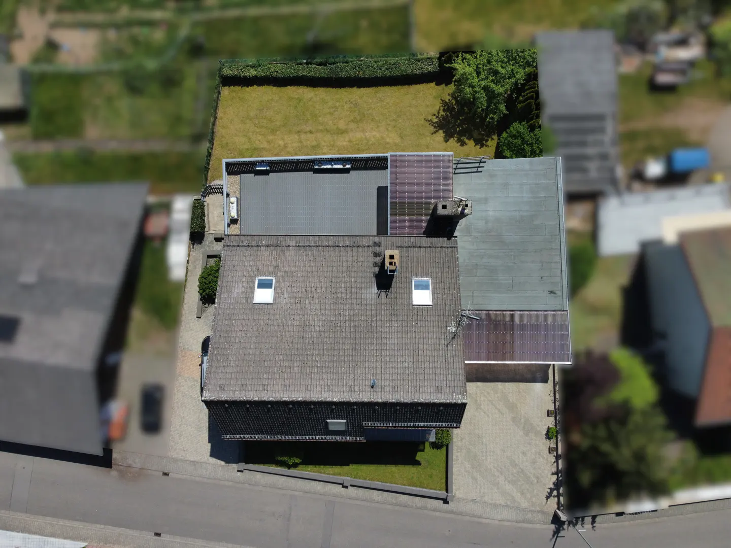Aerial view of a gray-roofed house with skylights, a green lawn, and solar panels on an adjacent flat roof.