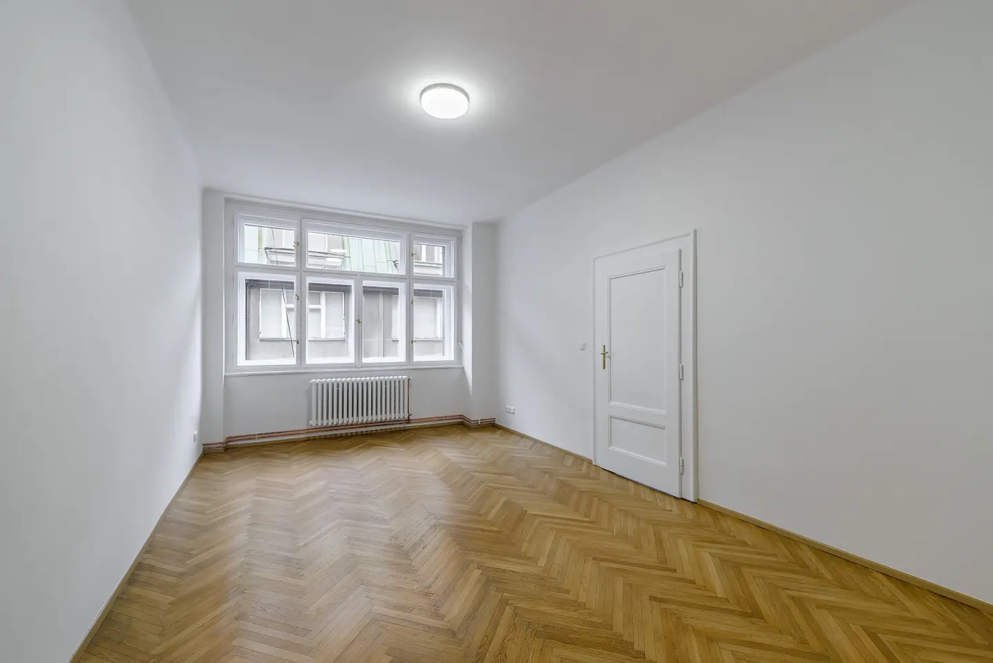 Bright, empty room with white walls, herringbone wood floor, large window, radiator, and a white door.