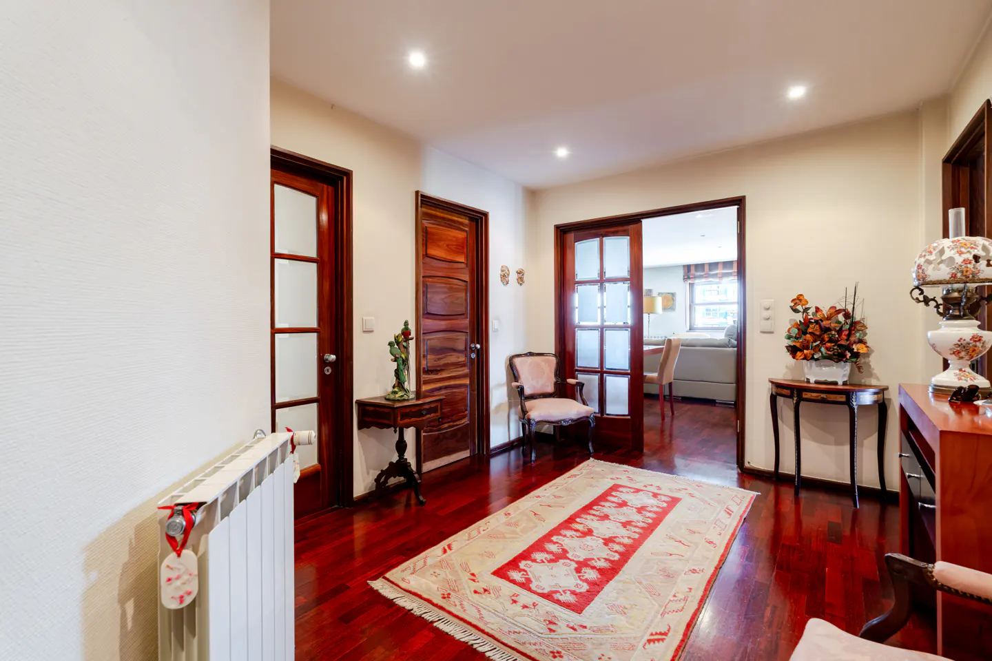 Bright hallway with dark wood floors, a red patterned rug, wooden doors, and elegant furniture under recessed lighting.