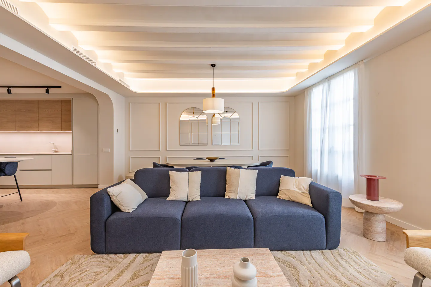 Living room with blue sofa, white pillows, and a marble coffee table on a patterned rug. Kitchen and dining area in the background.