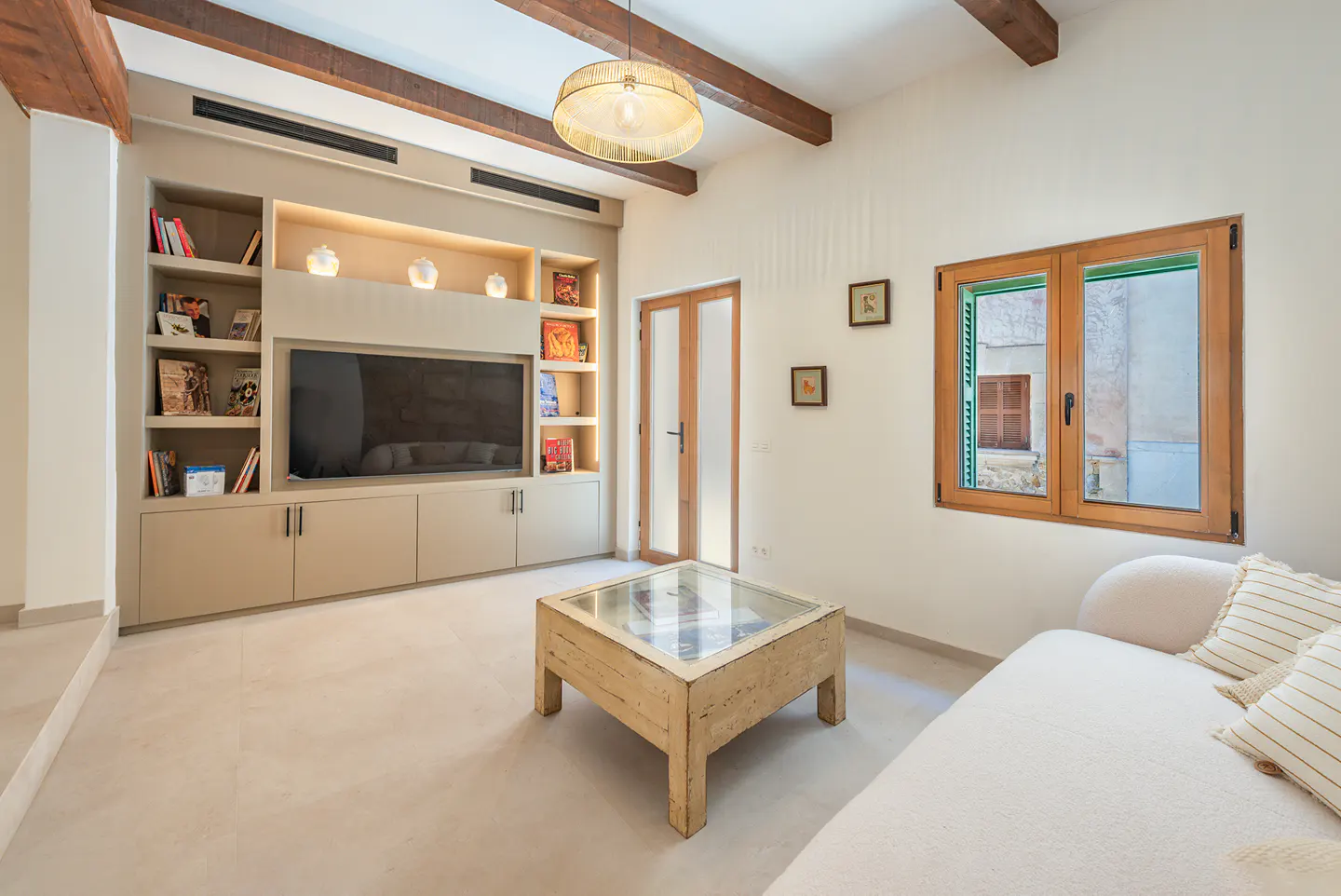 Living room with built-in shelves, TV, and cabinets. A wooden coffee table sits in front of a white sofa. A window with green shutters is on the right.