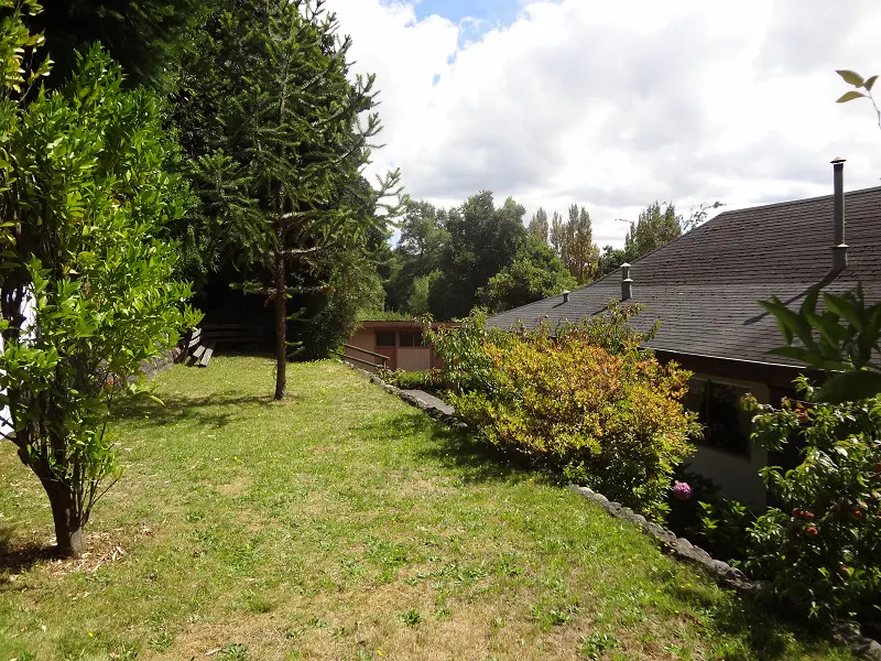 Lush green lawn with trees and shrubs, leading to a house with a dark roof under a partly cloudy sky.