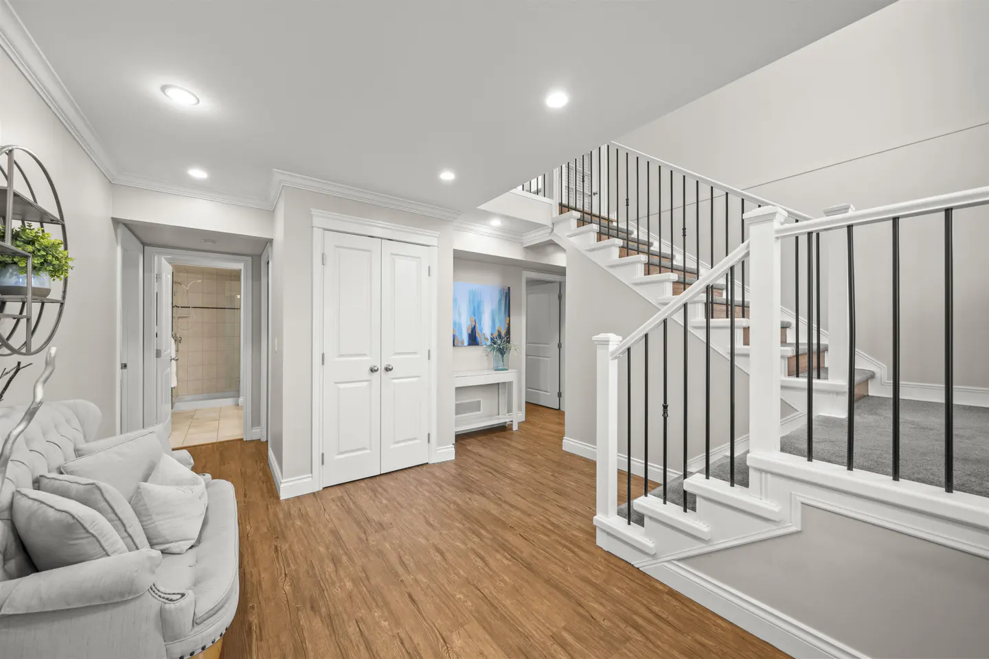 Bright foyer with wood floors, white walls, and staircase with black railings. A gray sofa sits near a bathroom door.