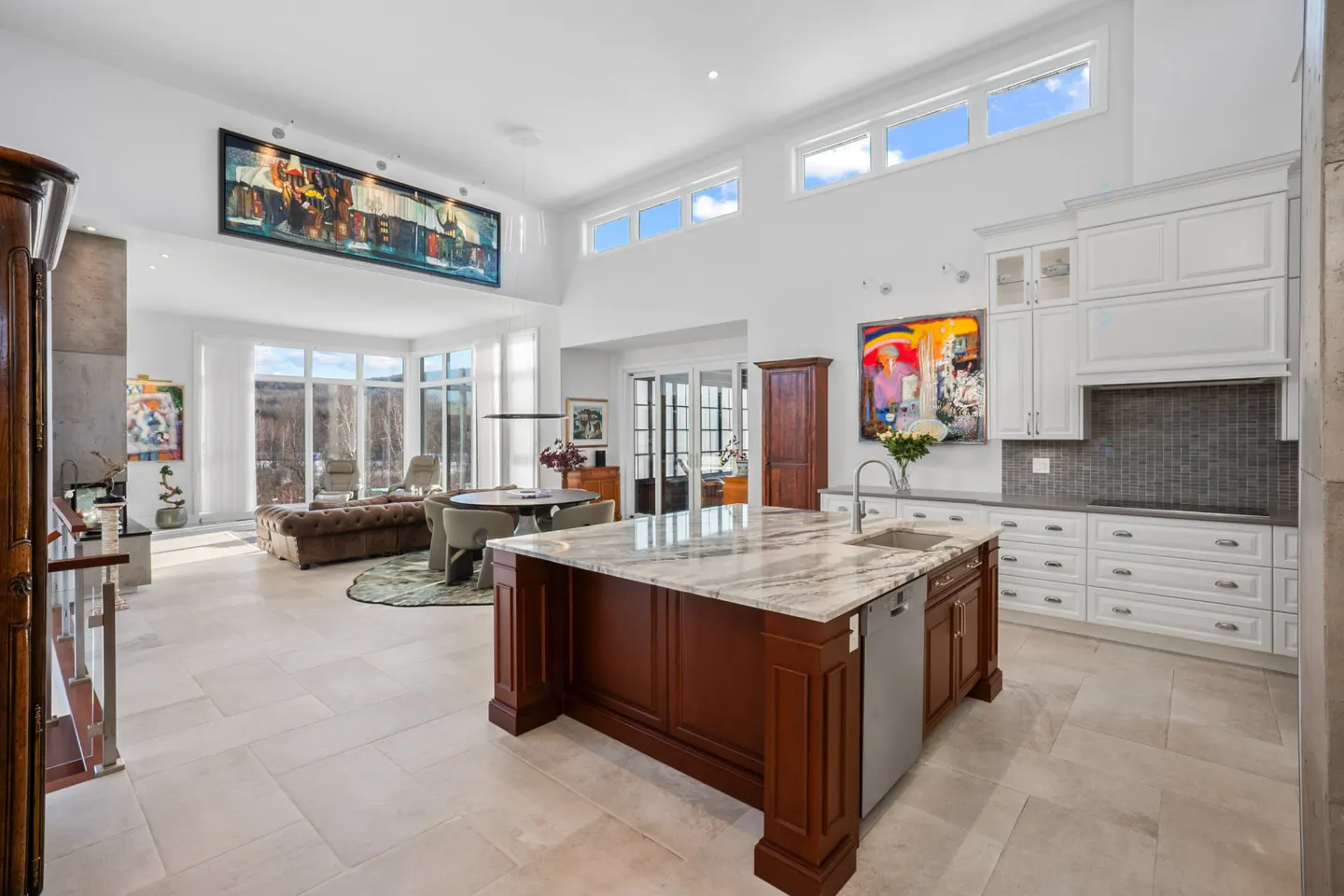 Bright, open-concept kitchen and living room with white walls, marble island, and large windows overlooking a wooded landscape.