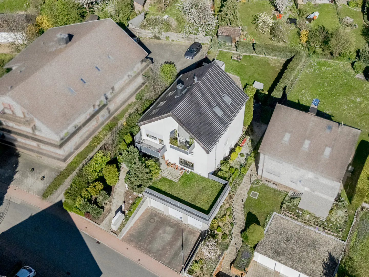 Aerial view of a white two-story house with a dark gray roof, surrounded by green lawns and trees. A car is parked in the driveway.