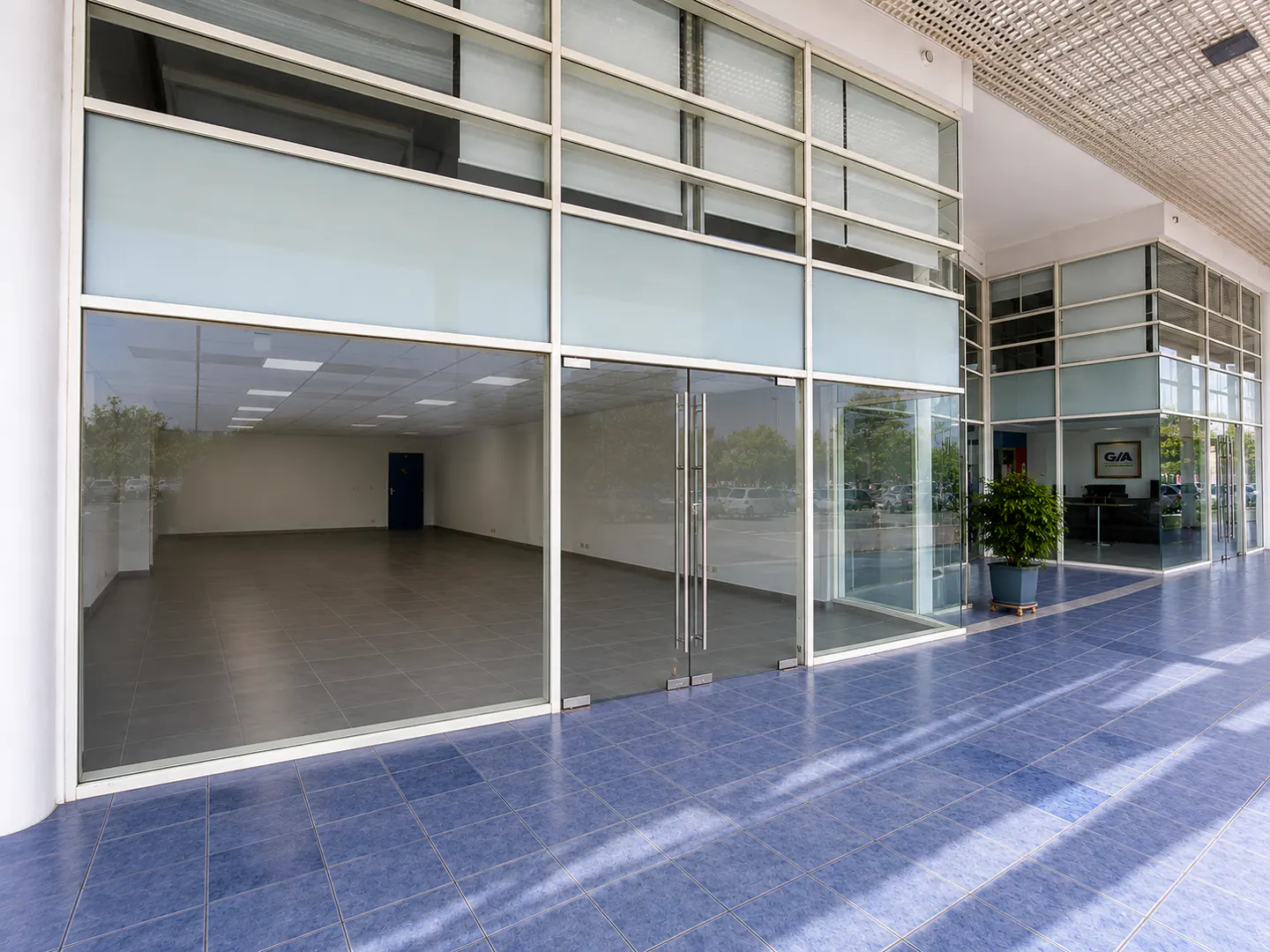 Empty commercial storefront with large glass windows, double glass doors, and blue tiled flooring under a covered walkway.