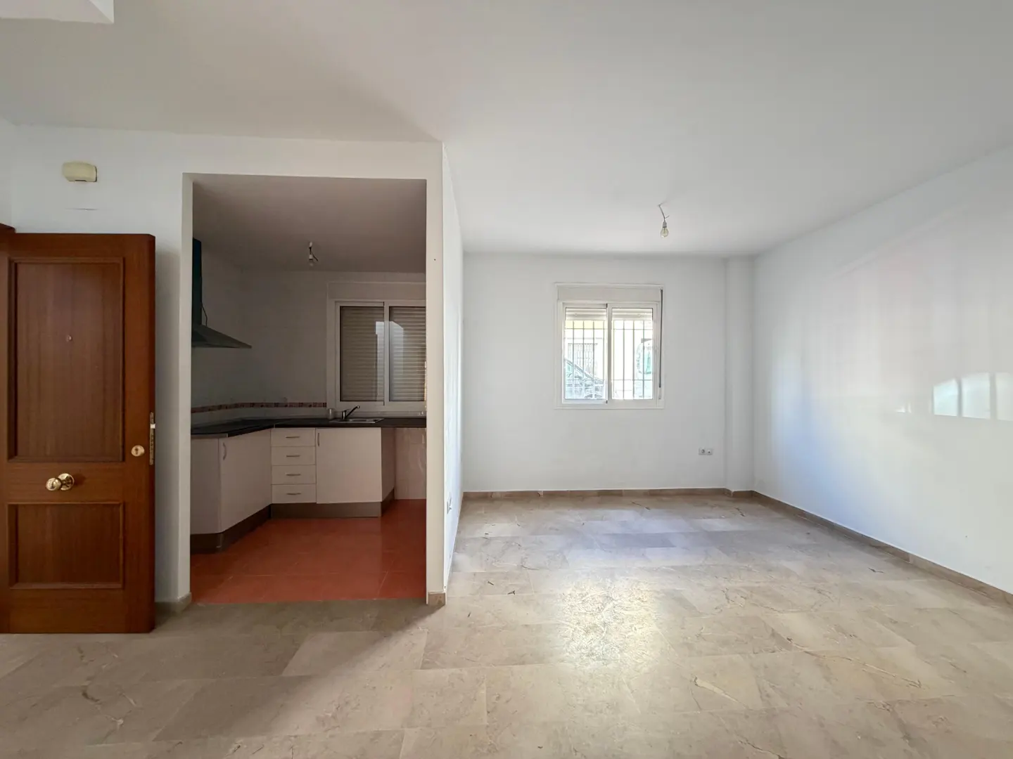 Bright, empty room with beige tile floor, white walls, and a window. An open doorway leads to a kitchen with white cabinets and a stainless steel range hood. A brown door is on the left.