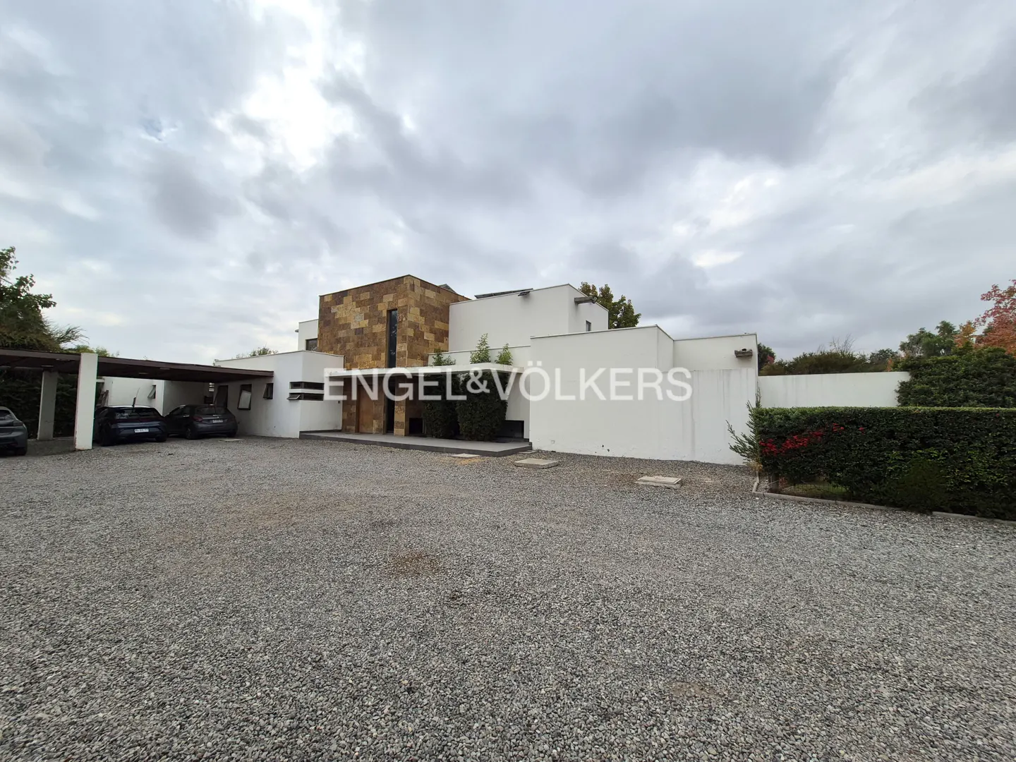 Modern white house with a stone accent wall and a gravel driveway under a cloudy sky. Cars are parked under a carport.