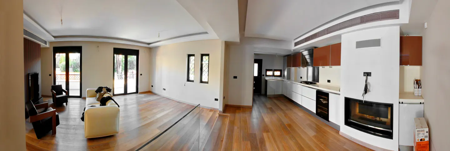 A panoramic view of a modern home interior with a living room and kitchen. The living room has a white sofa, dark chairs, and large windows. The kitchen features white cabinets and a fireplace.