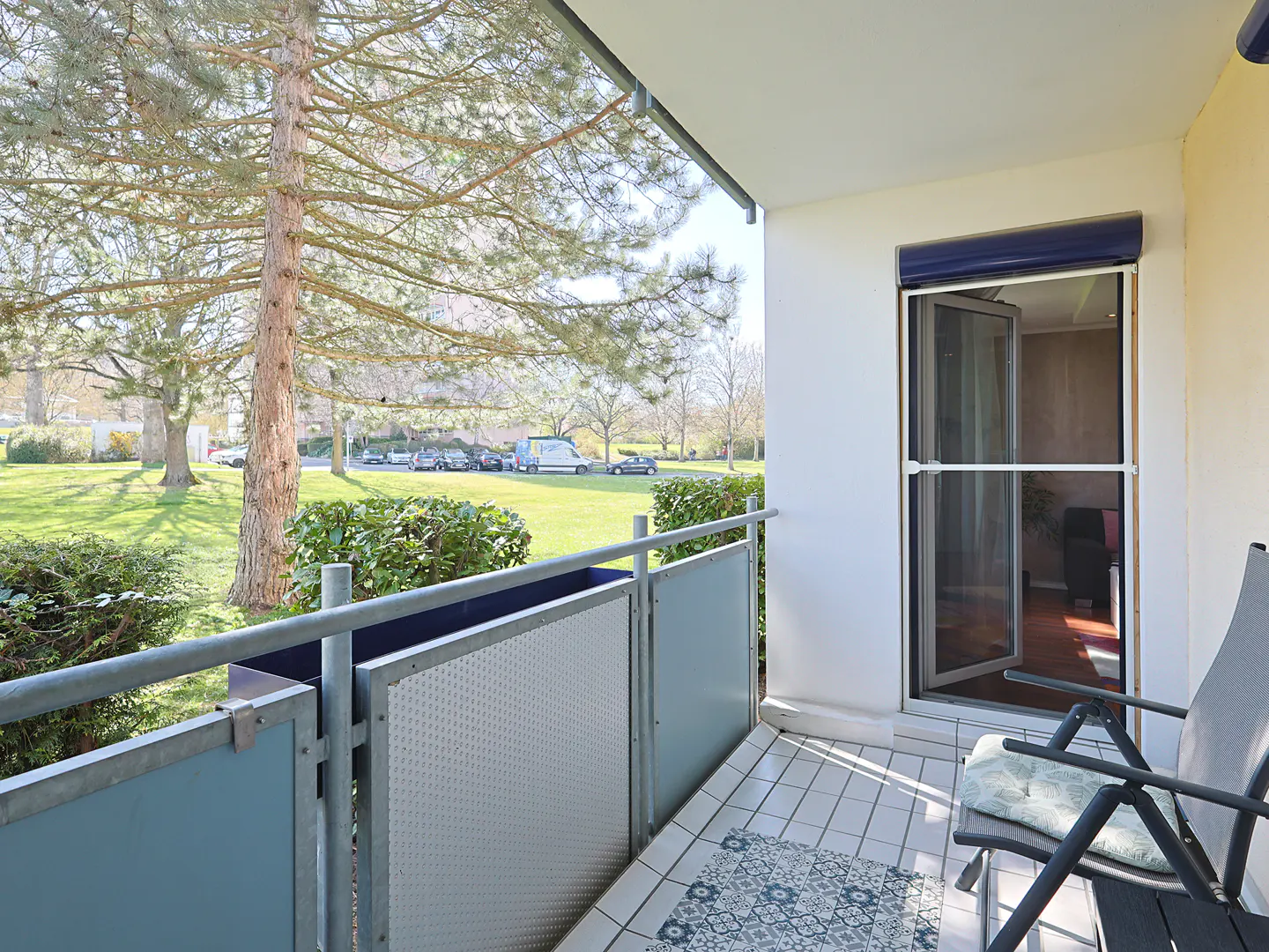 Balcony view with a gray chair, patterned tile rug, and metal railing overlooking a green lawn and trees.
