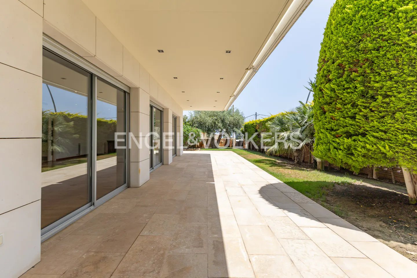 Covered patio with beige stone tiles, sliding glass doors, and green landscaping. Engel & Völkers logo visible.