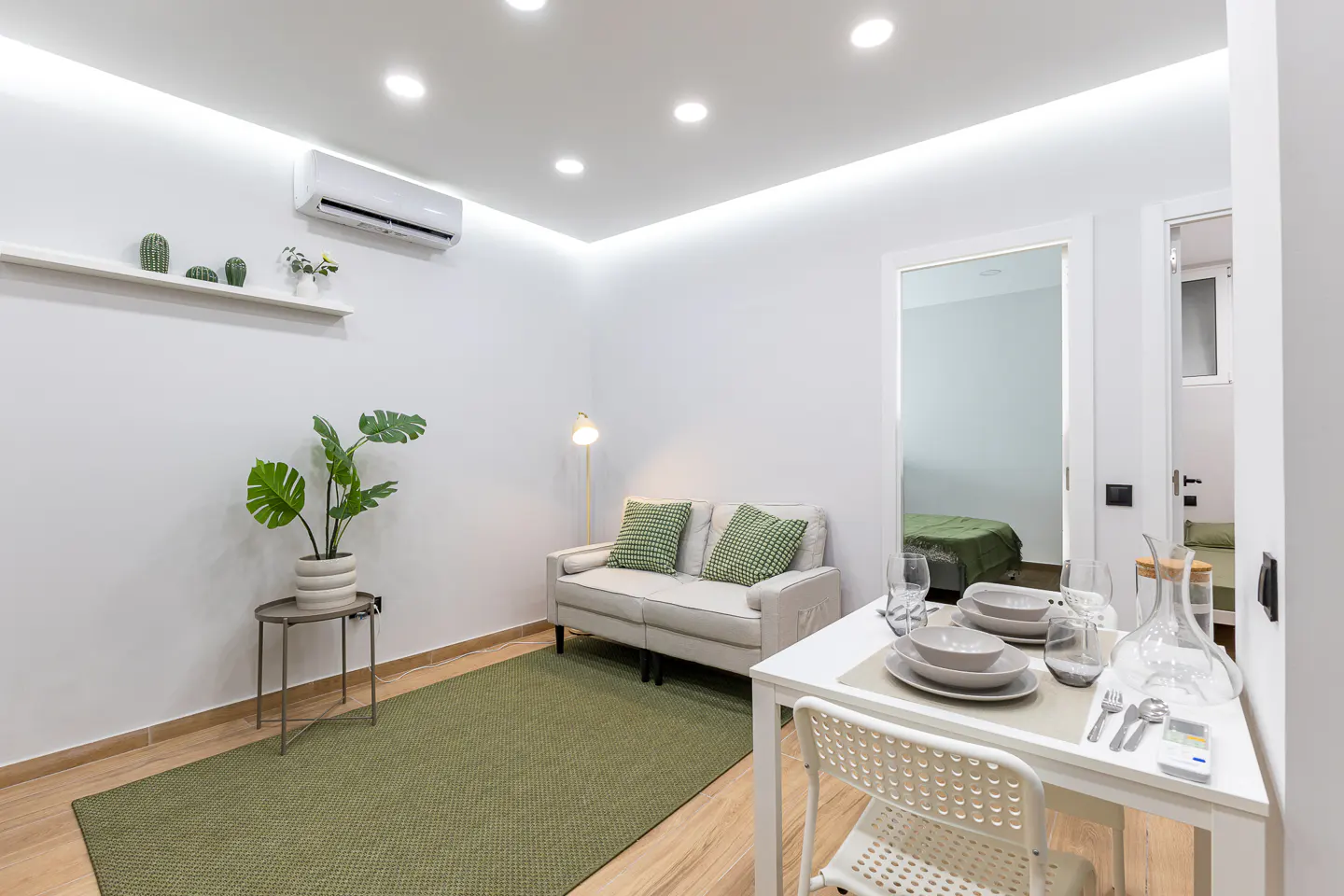 Bright, modern apartment interior with white walls, a green rug, and a small dining table set for a meal. A sofa and plant add a touch of home.
