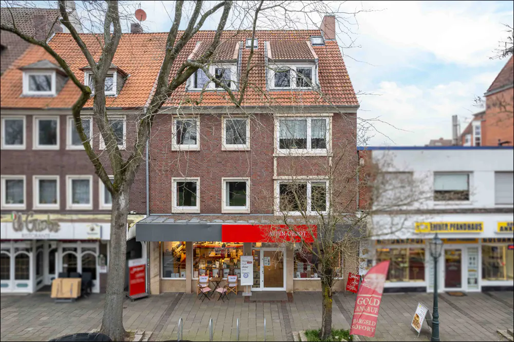 Three-story brick building with a red tile roof and a storefront on the ground floor. Trees frame the building.