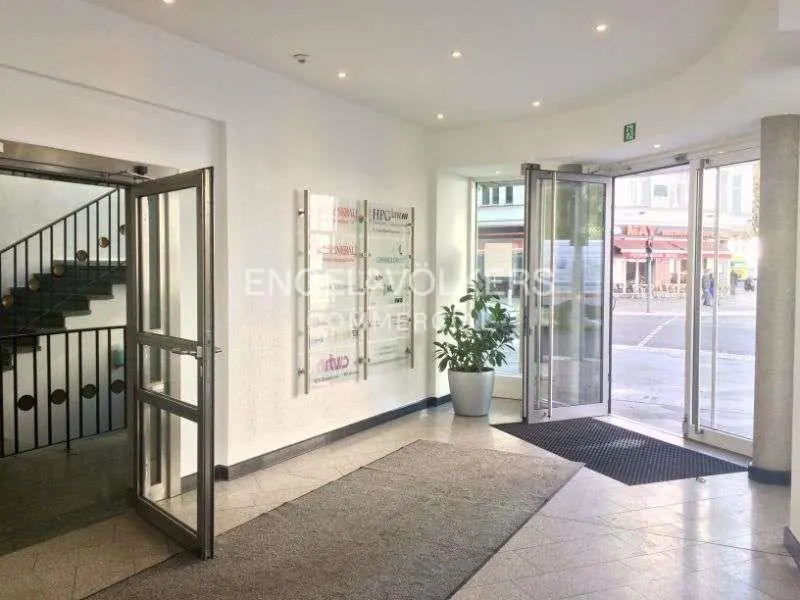 The Engel & Völkers office entrance features glass doors, a staircase, and a directory of businesses. A potted plant adds a touch of greenery.