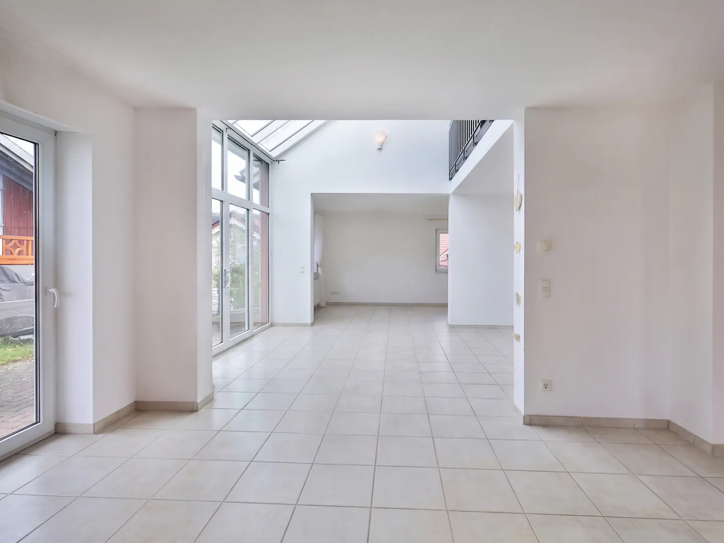Bright, empty room with white walls and tile floor. Large windows and a skylight provide natural light. A partial view of a second floor is visible.