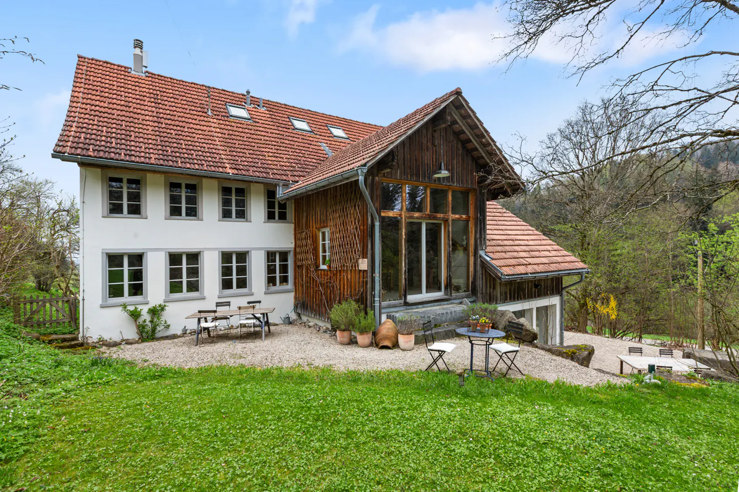 Exterior of a two-story house with a red tile roof, white walls, and a wooden extension, surrounded by green grass and trees.
