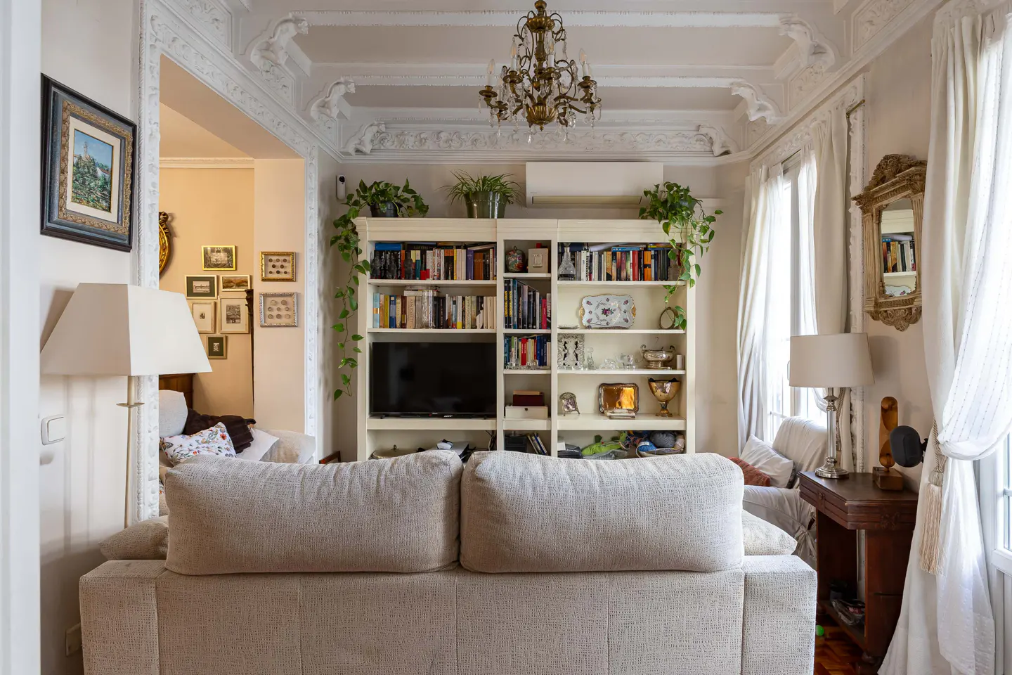 Living room with a beige sofa, a white bookcase with books and a TV, and a chandelier.