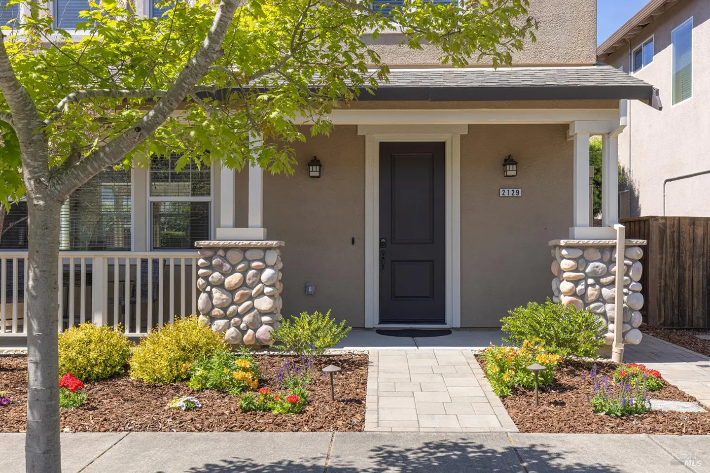 Front exterior of a tan house with a dark gray door, stone accents, and colorful landscaping.
