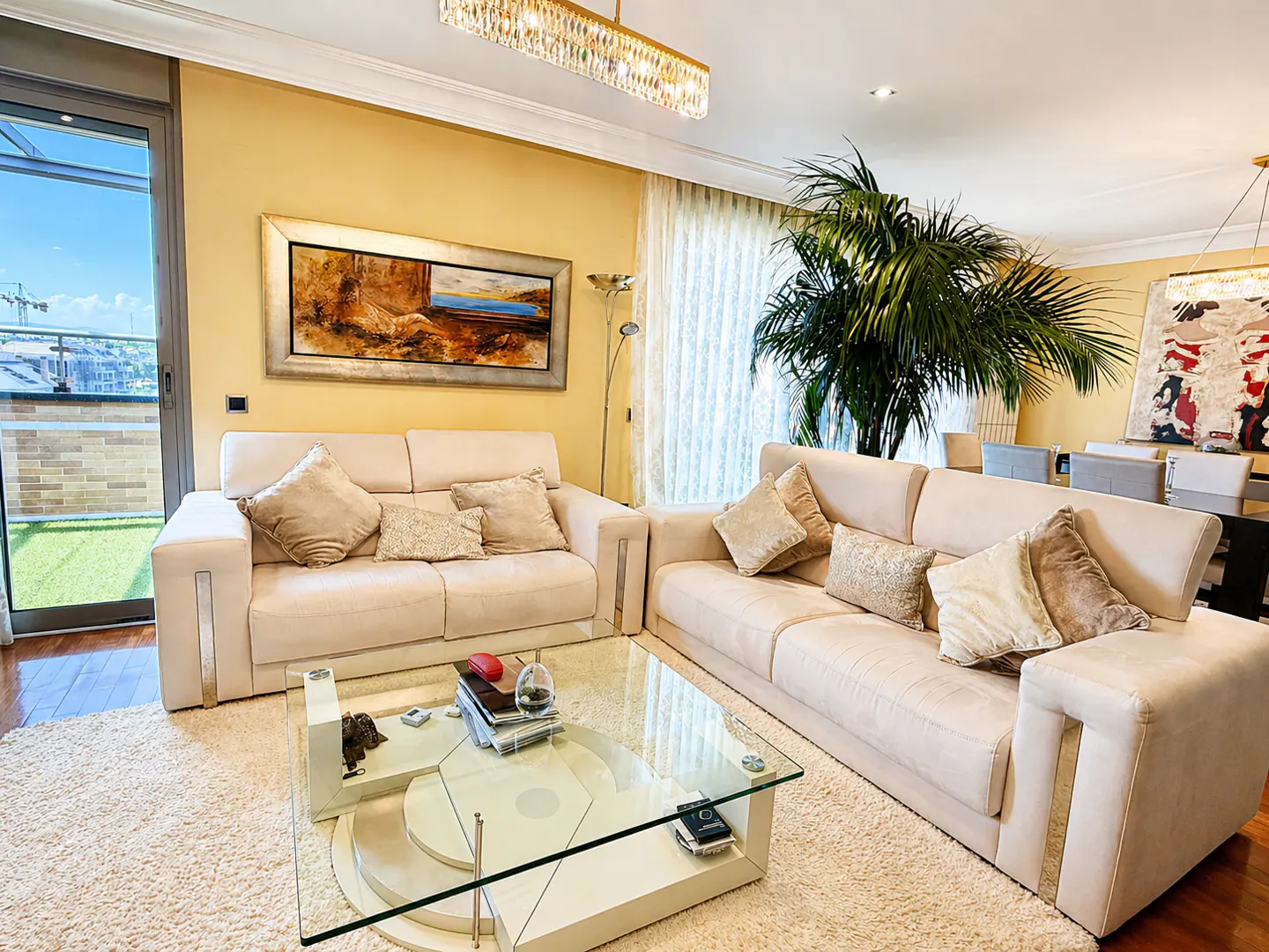 Living room with beige sofas, pillows, and a glass coffee table on a white rug. A painting hangs above the sofa, and a palm tree stands in the corner.