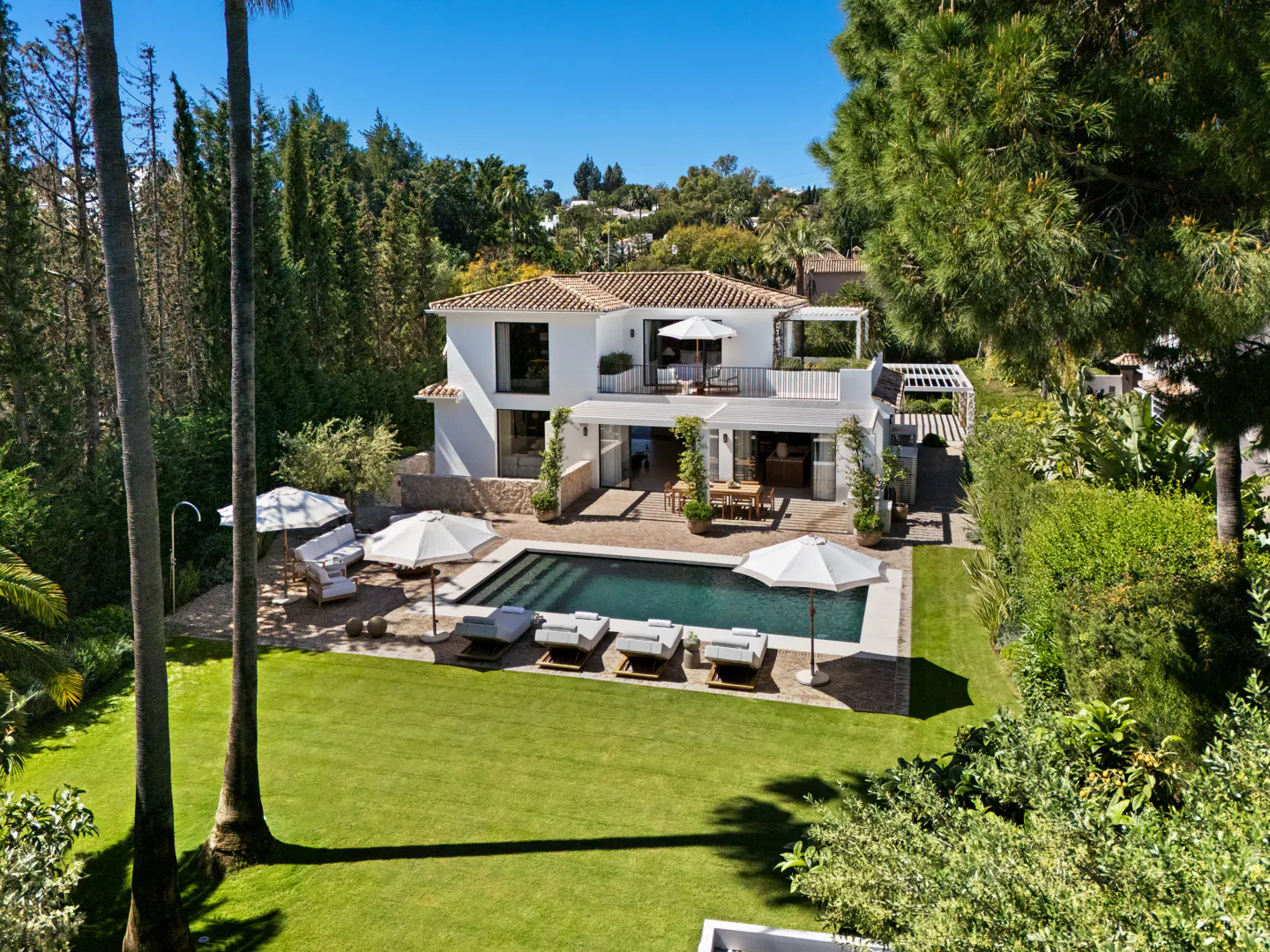 White villa with a pool, lounge chairs, and umbrellas in a lush, sunny garden setting.