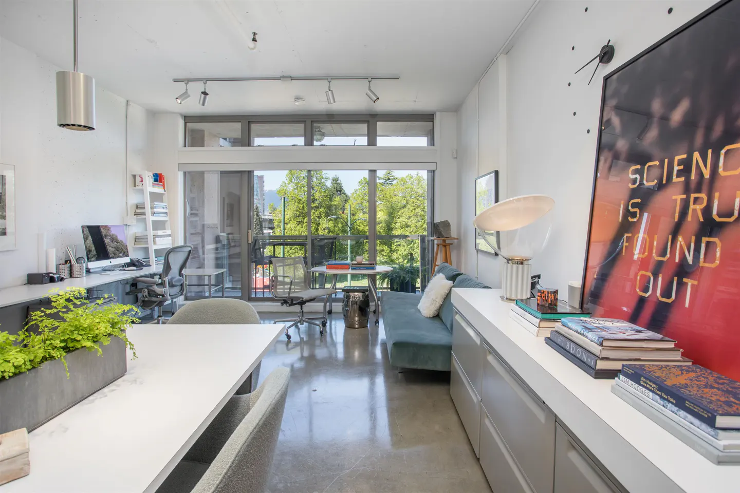 Bright, modern condo interior with a desk, blue sofa, and balcony overlooking green trees. A large "Science is Truth" print hangs on the wall.