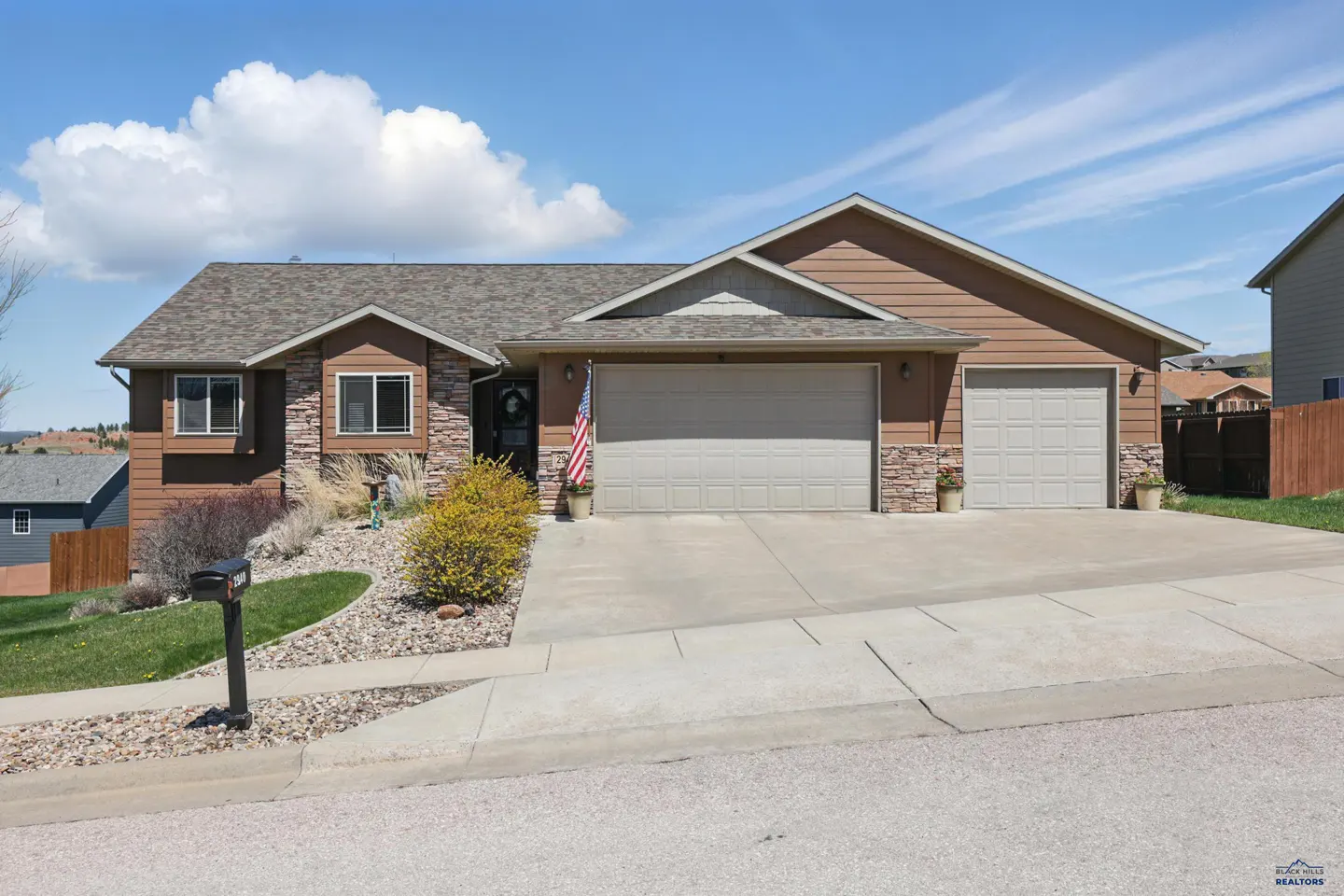 Single-story brown house with stone accents, a three-car garage, and an American flag by the front door under blue sky.