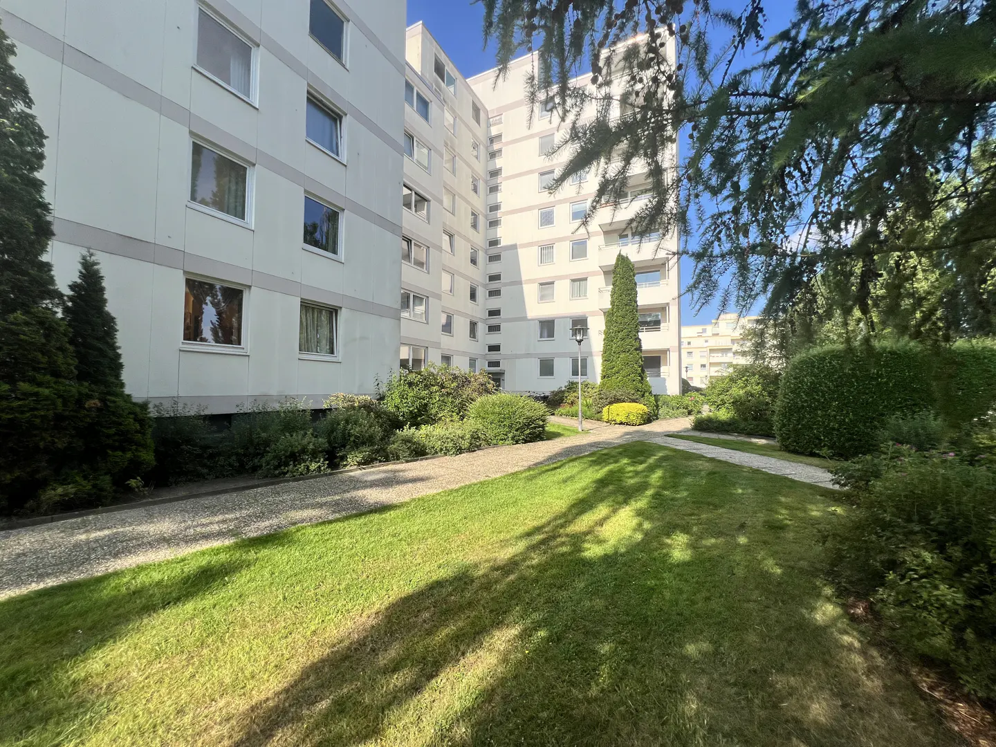 Exterior view of a tall, light gray apartment building with green lawn, trees, and gravel path on a sunny day.