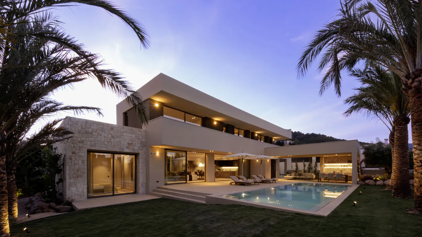 Modern beige house with a pool at dusk. Palm trees frame the view of the two-story home with outdoor seating and lighting.