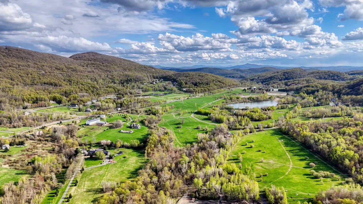 Aerial view of a lush green valley with rolling hills, scattered houses, and a small lake under a partly cloudy blue sky.