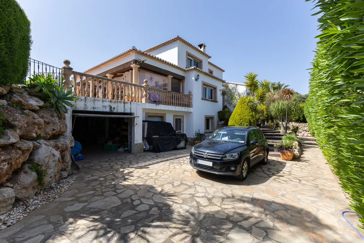 Two-story white house with a balcony, garage, and a black car parked on a stone driveway. Green hedges and trees surround the property.
