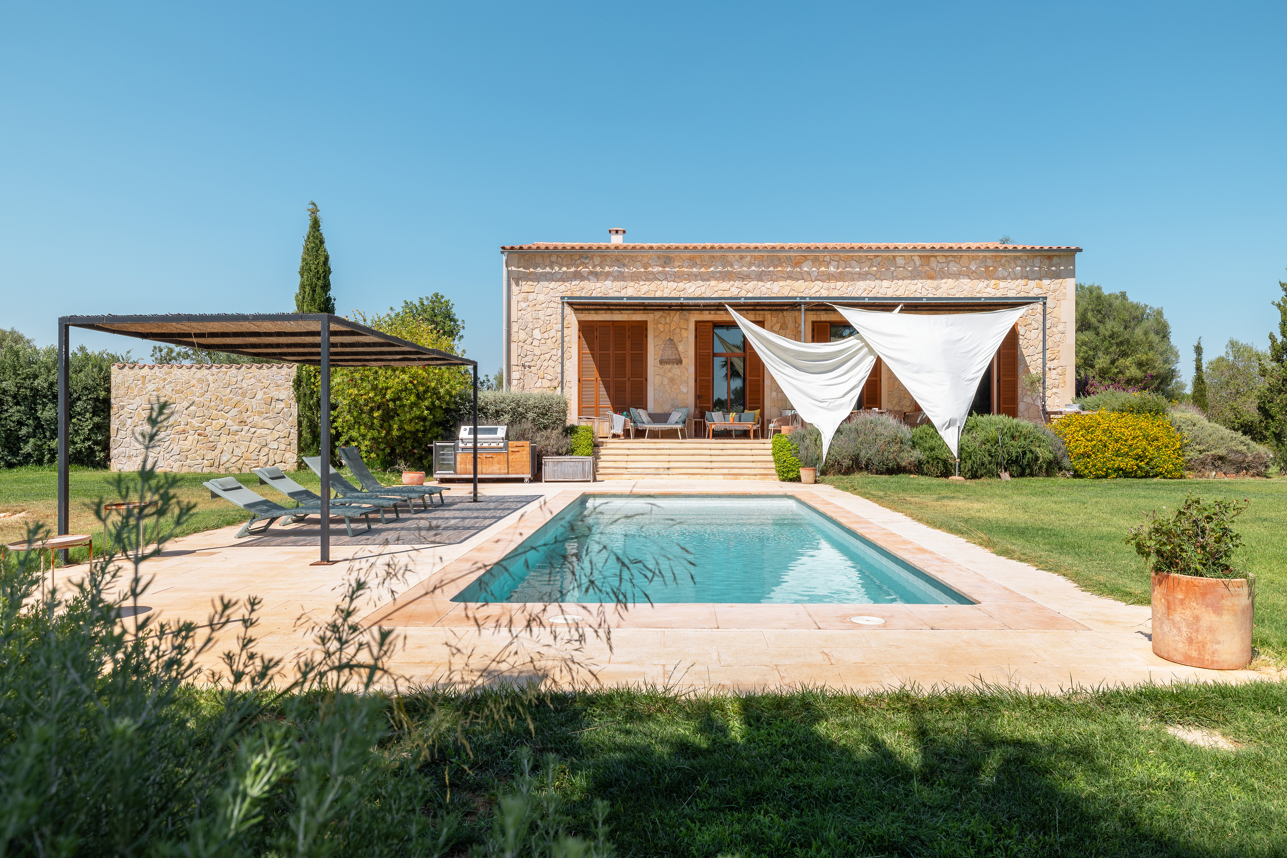 Stone villa with a swimming pool, sun loungers under a pergola, and white shade sails on a sunny day. [AI-generated text]