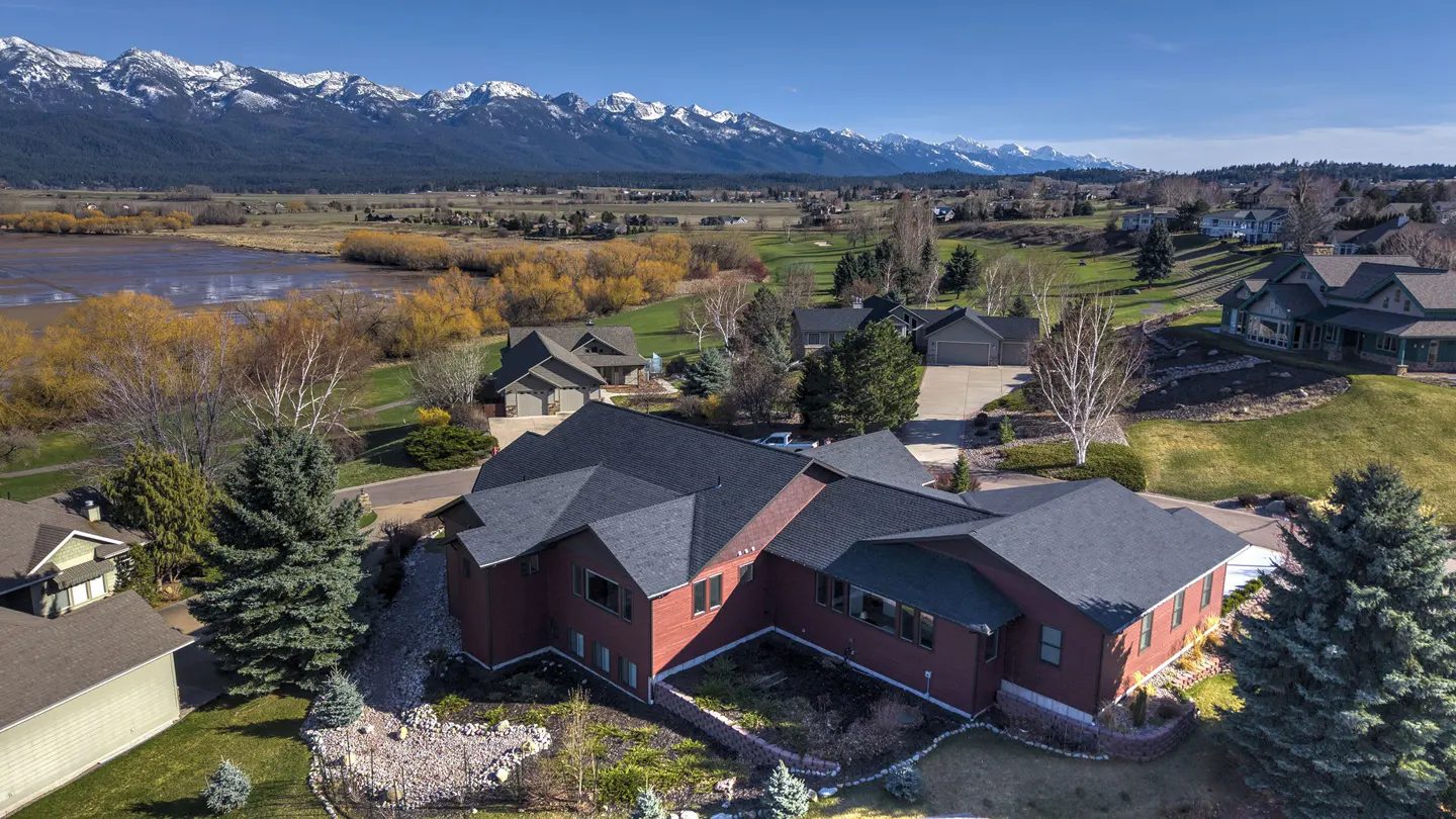 Aerial view of a red house with a dark roof, green lawn, and snow-capped mountains in the background.