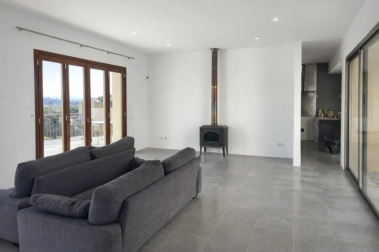 A bright, modern living room with a gray sofa, wood-burning stove, and folding glass doors to a balcony.