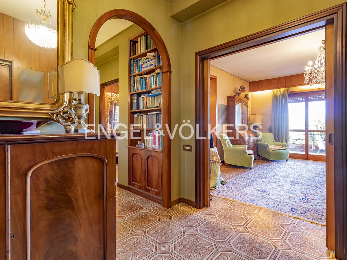 Hallway with patterned tile floors, a wooden arched bookshelf, and a view into a living room with green armchairs.