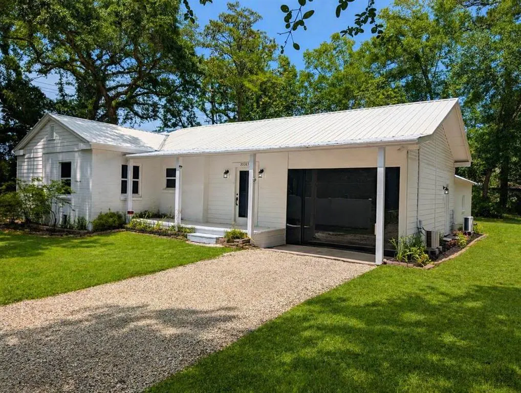 Exterior of a white, one-story house with a metal roof, black garage door, gravel driveway, and green lawn.