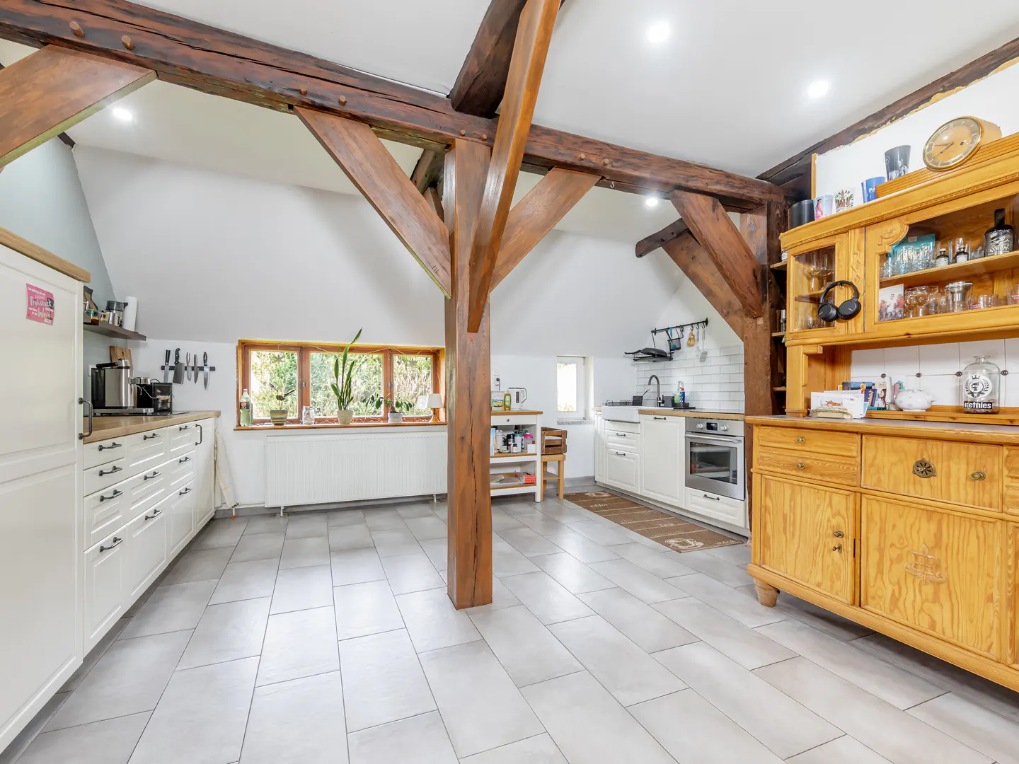 Bright kitchen with white cabinets, gray tile floor, and exposed wooden beams. A large wooden hutch stands against the wall.