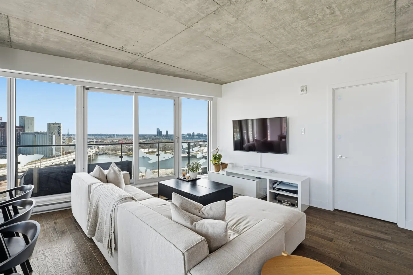 Bright living room with a white sectional sofa, black coffee table, and a large window with a city view. TV on the wall.