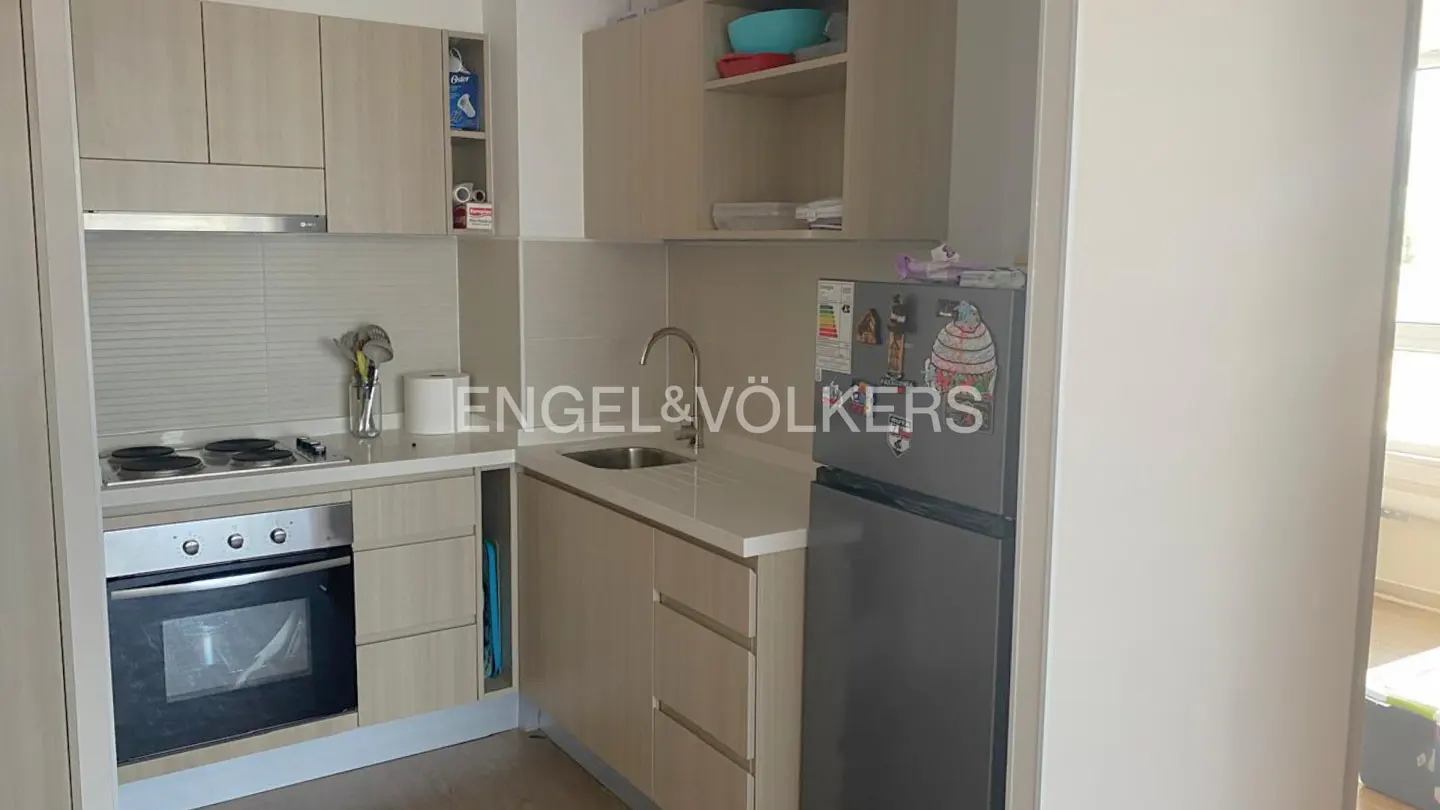 A compact kitchen features light wood cabinets, a stainless steel oven and cooktop, and a gray refrigerator with magnets.