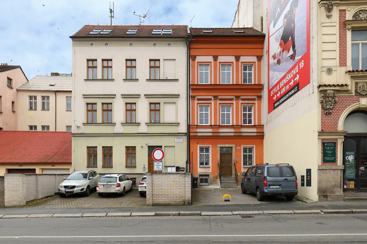 A street view of a light-colored building next to a narrow orange building with parked cars in front.