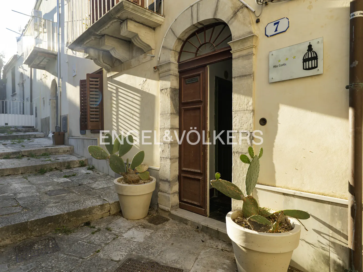 Entrance to a stone building with a wooden door, two potted cactus plants, and a house number 7 sign on a sunny street.
