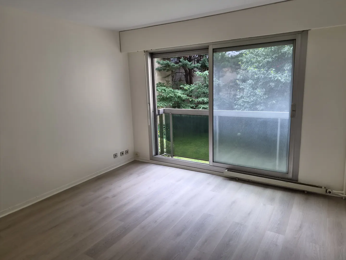 Empty room with light wood-look flooring, white walls, and a sliding glass door opening to a balcony with green trees.