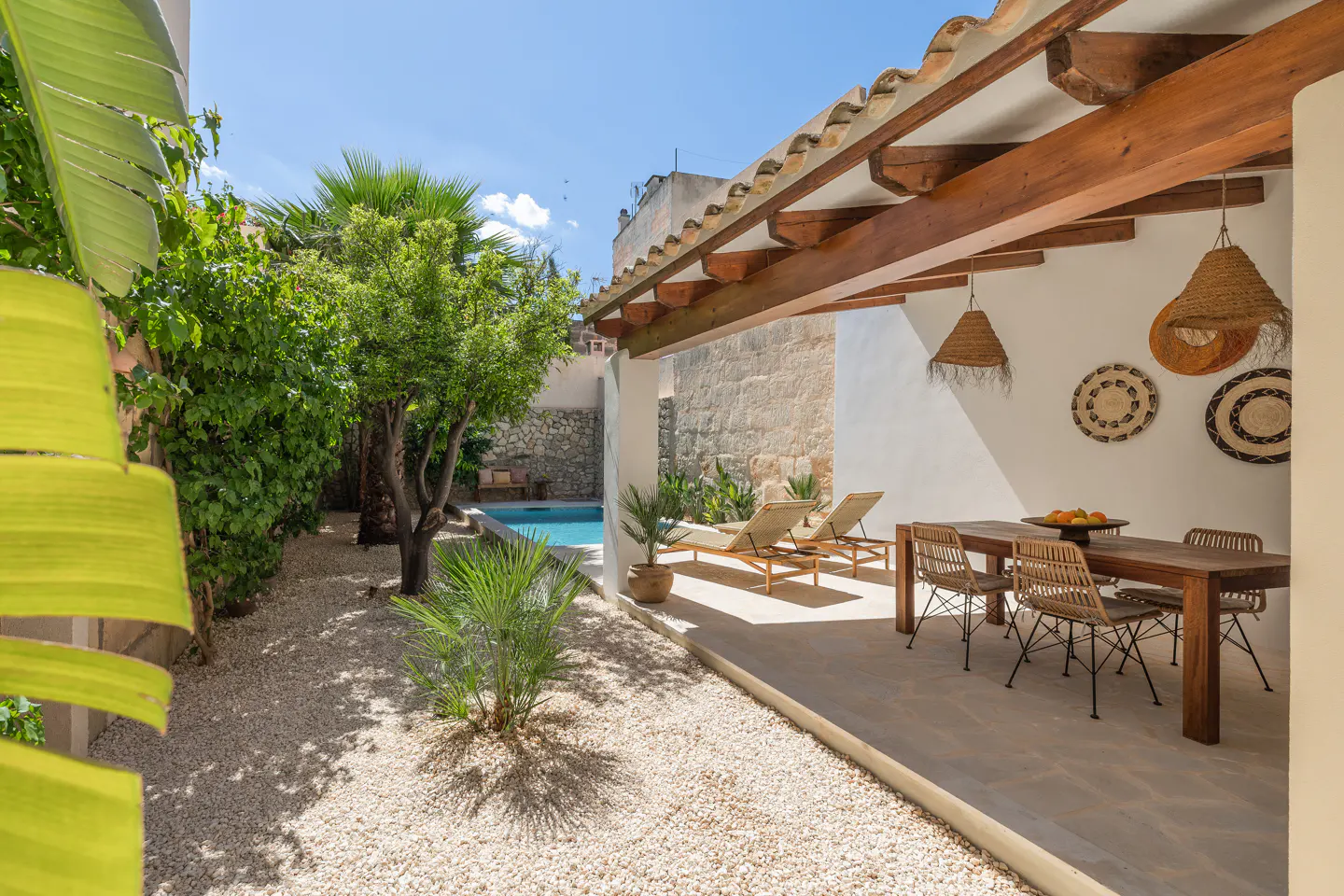 Outdoor patio with a wooden table, chairs, and lounge chairs near a pool, surrounded by greenery and white gravel.