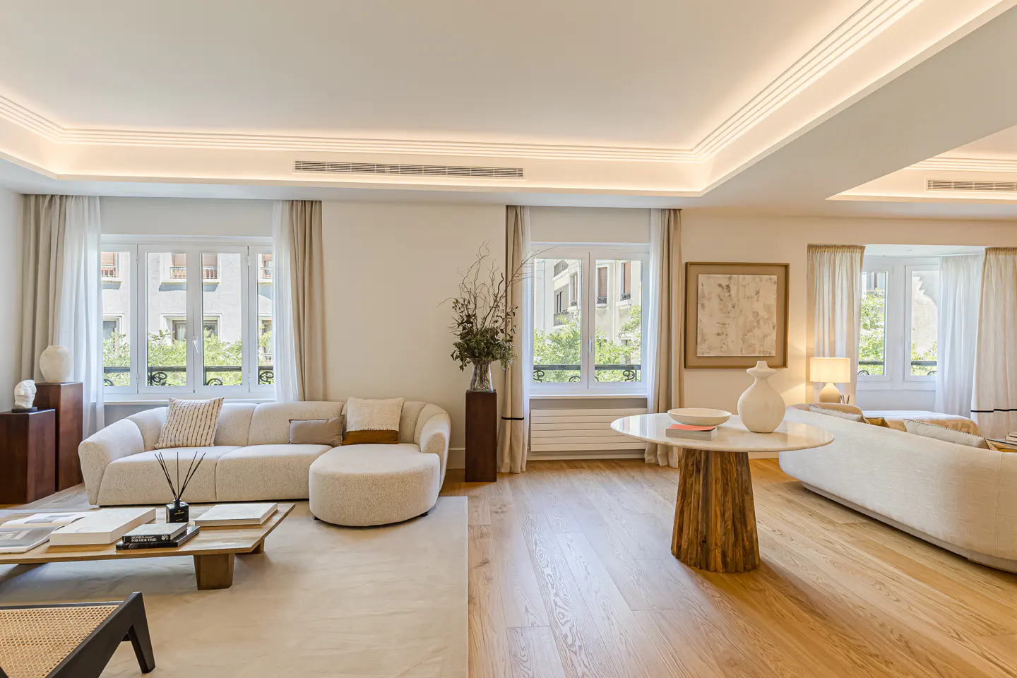 Bright, modern living room with a cream sofa, wooden coffee table, round pedestal table, and large windows with curtains.