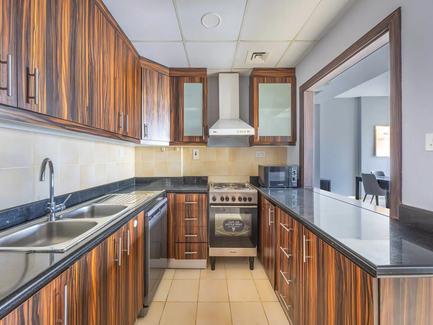 A kitchen with wood cabinets, black countertops, stainless steel sink, and appliances. A pass-through to the dining area is visible.