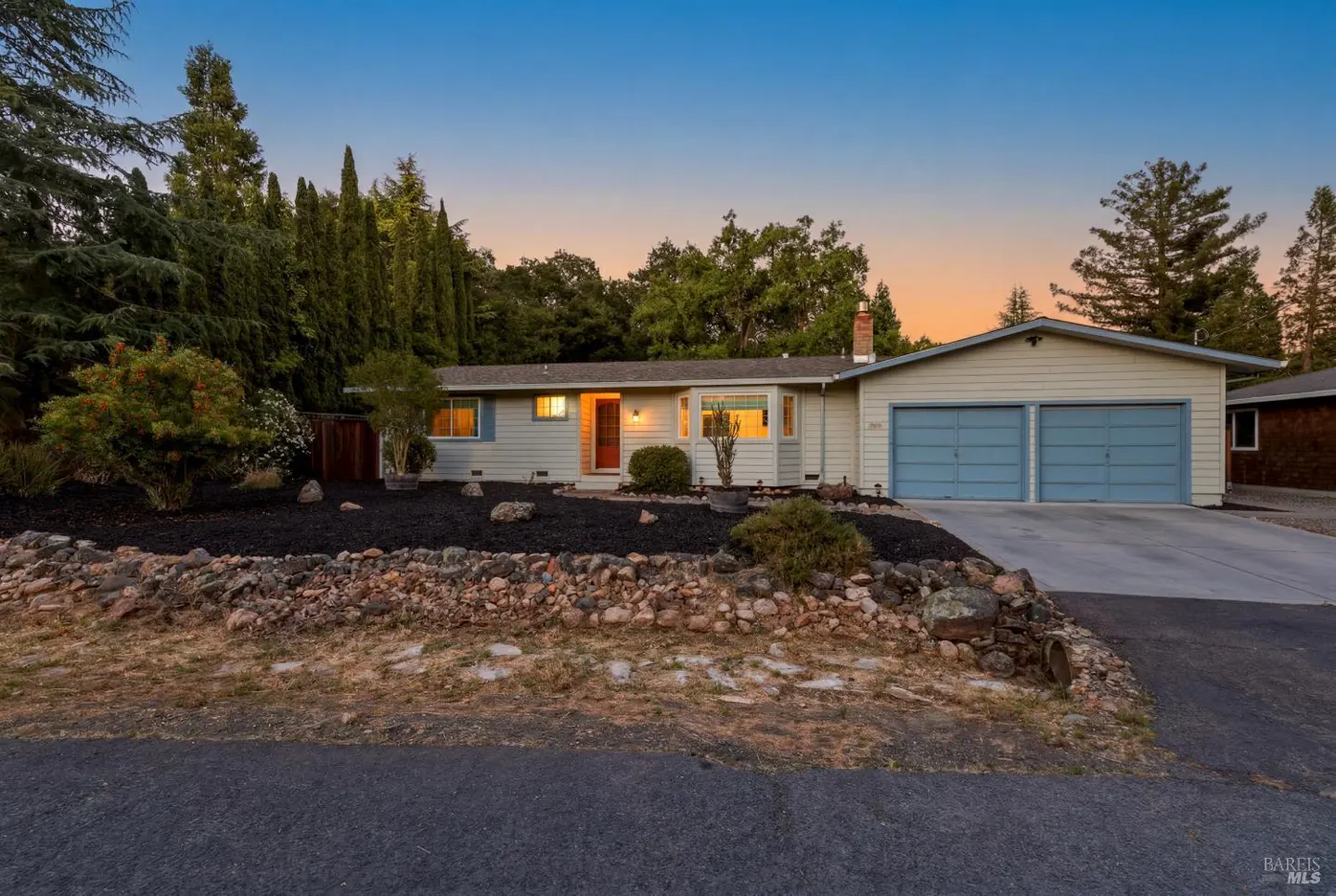 Single-story home with light siding, two-car garage, and a rock-walled front yard at sunset.