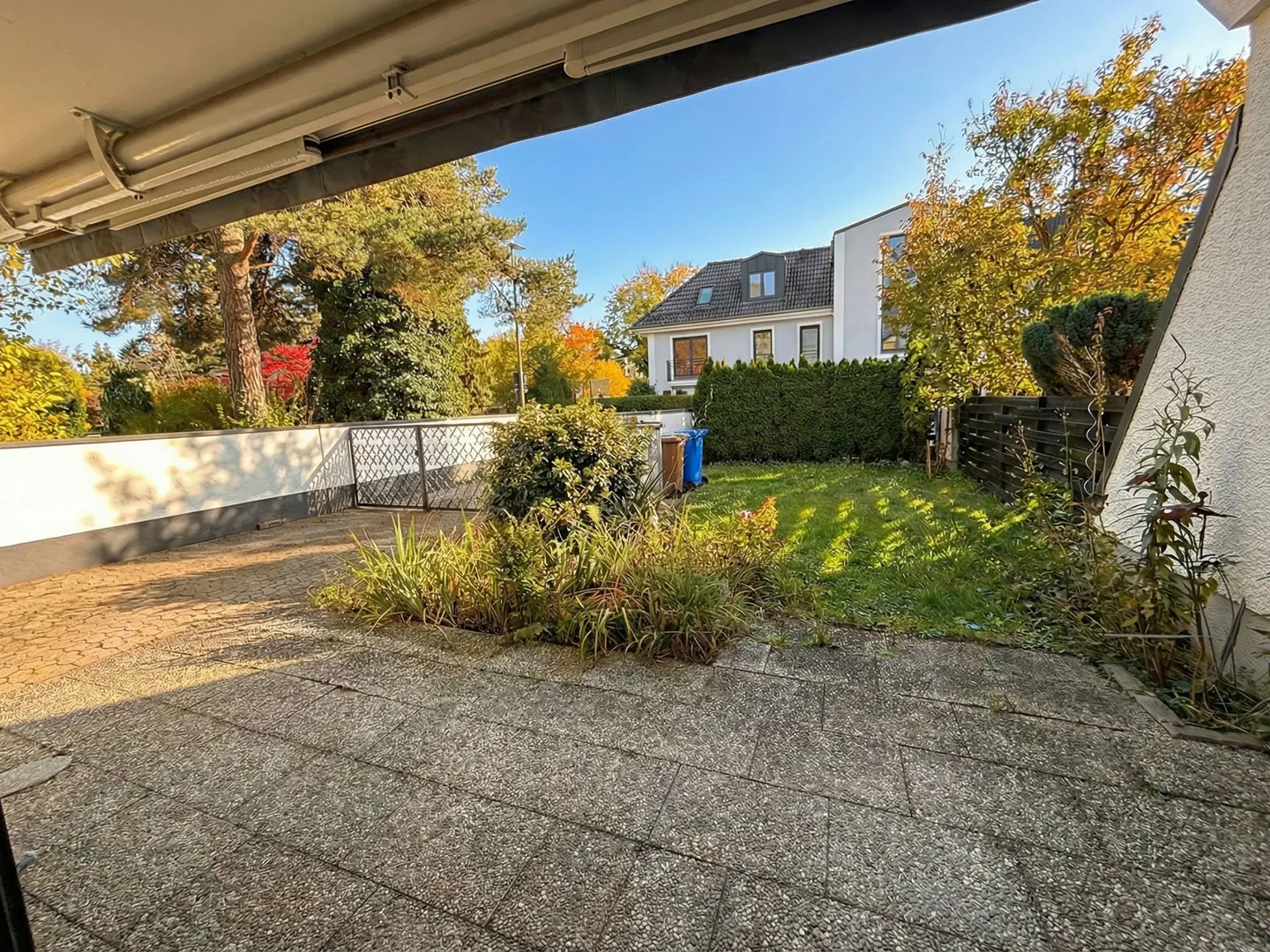 View from a covered patio to a yard with grass, plants, and a white house in the background on a sunny day.