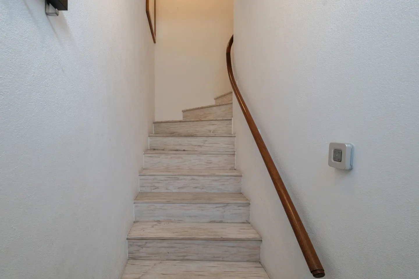Interior view of marble stairs with a wooden handrail against a white wall.
