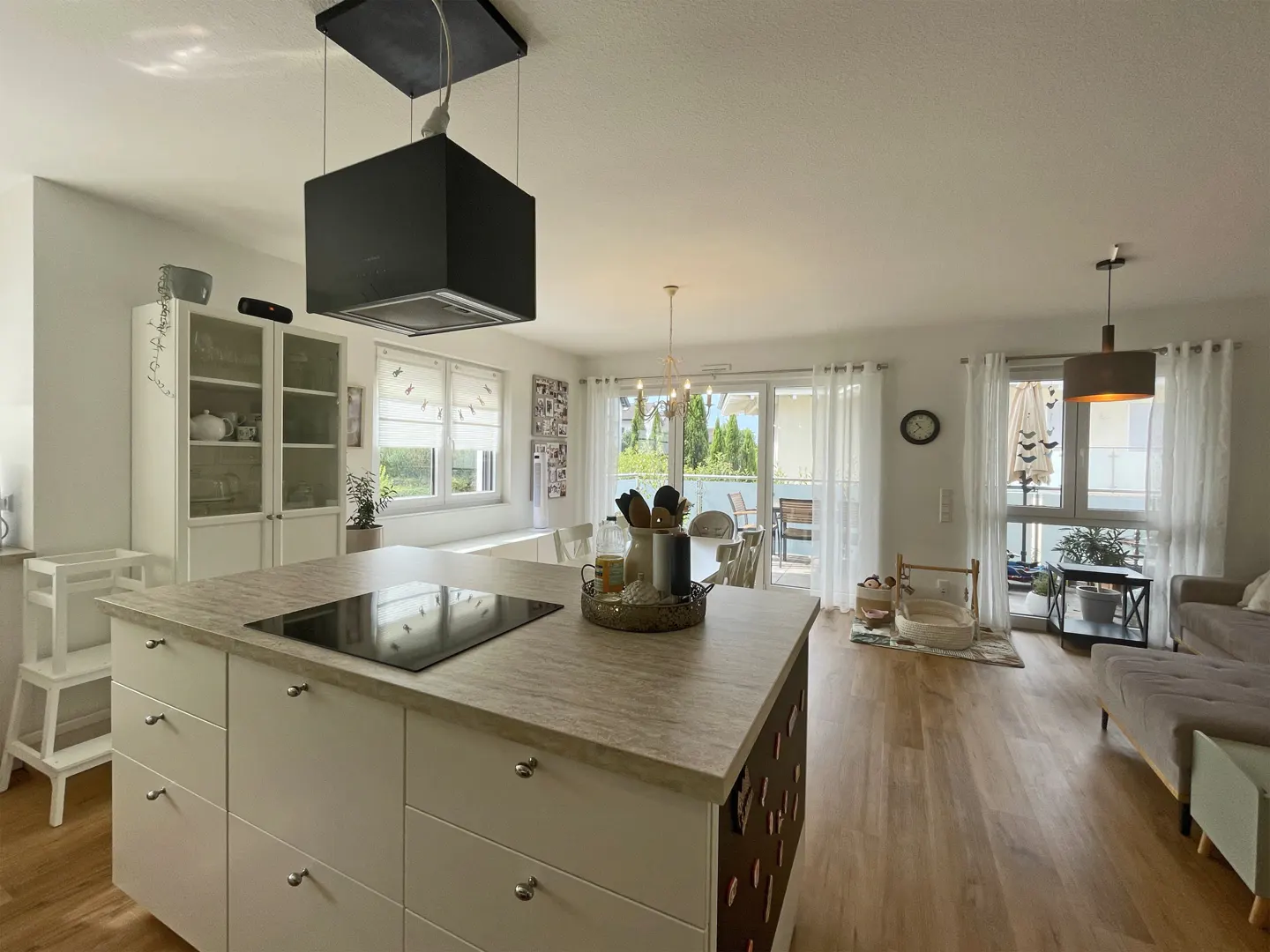 Open-concept kitchen and living area with white cabinets, island, and wood floors. Natural light from windows and balcony doors.